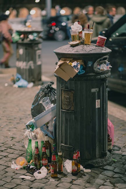 A black public rubbish bin situated on a cobblestone street, over which several discarded paper food containers, plastic drink cups, and napkins are scattered. The lid of the bin is open, with some trash protruding from it, including a cardboard box and a crumpled plastic bag. Several empty glass bottles, mostly beer bottles with green and brown tint, are placed around the base of the bin. On top of the bin, there are a few used paper cups with plastic lids, some with contents visible, and sprinkled paper tissues. In the background, blurred figures of pedestrians and parked cars are visible, indicating an urban outdoor setting. A large, dark-colored waste collection bag is leaning against the side of the bin. The scene depicts typical street littering and highlights the importance of proper waste disposal, which could be addressed through private rubbish removal services like those offered by Rubbish Clearance Enfield, to promote cleaner public spaces.