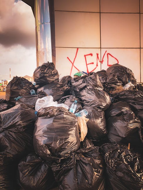 A large pile of black plastic rubbish bags filled with waste is stacked against a modern building with a tiled exterior wall, partially covered by graffiti spelling 'XEND' in red spray paint. Several clear plastic water bottles are visible protruding from some of the bags. The scene appears to be outdoors, on a paved surface, with the sky showing a mix of clouds and sunlight in the background. The rubbish bags are tightly packed and piled high, suggesting an instance of excess waste awaiting removal or disposal. The image relates to private rubbish collection services offered by Rubbish Clearance Enfield, highlighting the need for on-site waste clearance and alternative methods of rubbish removal outside of local authority collection schedules.