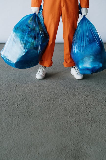 A person dressed in orange overalls and white sneakers is standing on a gray concrete surface, holding two large blue plastic bags filled with waste or rubbish, with the bags appearing semi-transparent and bulging from the contents. The background is plain and minimal, with no specific environmental details visible, emphasizing the focus on the bags and the individual’s lower body. This scene reflects an informal or private waste collection scenario, which Rubbish Clearance Enfield may facilitate as an alternative to standard municipal rubbish disposal, illustrating the process of rubbish removal or on-site clearance. The lighting is neutral, providing clear visibility of textures and colors, suitable for describing waste handling or rubbish removal services associated with bulky waste clearance in Enfield.