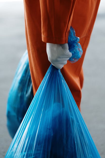 A close-up of a person wearing an orange protective jumpsuit and white gloves, holding a tightly knotted blue plastic rubbish bag in their right hand. The bag appears to be filled with waste materials, showing a smooth, shiny surface with some creases and folds. The background is blurred, suggesting an outdoor environment, possibly near a waste collection or disposal site, with natural daylight illuminating the scene. The image emphasizes the safe handling of waste packaging, aligning with rubbish removal practices and supporting private waste management solutions such as those offered by Rubbish Clearance Enfield, particularly for bulky or general rubbish disposal tasks.