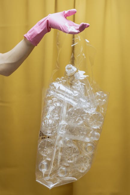 A person wearing a pink rubber glove is holding a clear plastic rubbish bag filled with used transparent plastic bottles and packaging, which appears to be ready for collection or recycling. The bag is large, tightly filled with various plastic waste items, and slightly crinkled, indicating it is full and ready for disposal. The background is a plain yellow curtain or wall, and the lighting is even, highlighting the shiny, smooth surface of the plastic waste. This scene is typical of waste management or rubbish removal activities, where plastics are gathered for recycling as part of an alternative waste handling method. The image emphasizes the importance of proper plastic waste separation and disposal, which Rubbish Clearance Enfield offers as part of their rubbish services in local areas, including private and on-site collection options for household waste.