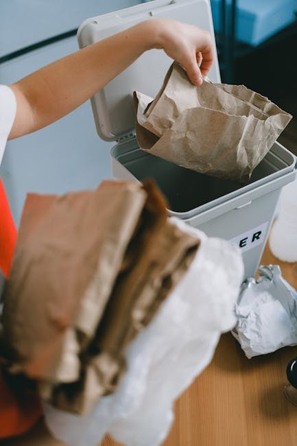 A person placing a crumpled brown paper bag into a small, rectangular, white bin designated for waste. The bin is positioned on a wooden surface, with other crumpled paper rubbish nearby. The environment appears to be indoors, possibly a home or office setting, with a neutral background. The paper bag has a slightly rough texture and light brown coloration, contrasting with the smooth, white surface of the waste bin. The scene reflects an act of private waste disposal, aligning with the context of rubbish removal services like those offered by Rubbish Clearance Enfield, illustrating the process of regular domestic or office rubbish collection as part of an alternative waste handling method.