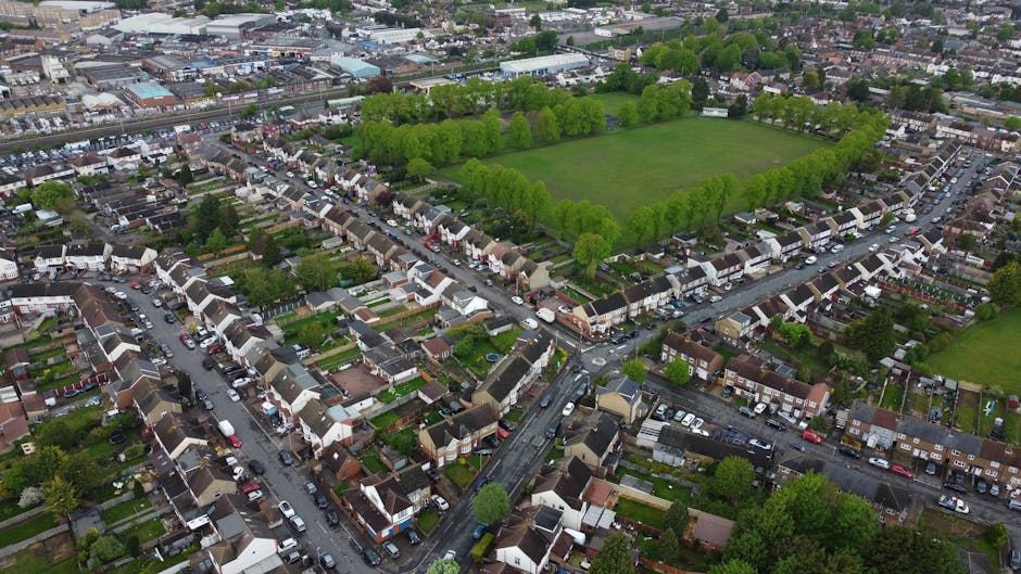 An aerial view of a residential neighbourhood featuring a large, green rectangular park surrounded by dense, mature trees lining its perimeter. The park is bordered by a mixture of terraced houses and detached homes with small gardens, paved pathways, and vehicle driveways visible along the streets. In the foreground, tightly packed rows of houses with rooftops of varying shades of brown, grey, and white create a uniform pattern, with cars parked along the narrow, winding roads. Some houses have visible front yards with hedges, lawns, and small fences. The area's environment appears calm and well-maintained, with greenery interspersed among built structures. In the background, more industrial or commercial buildings and additional residential areas spread outward, illustrating the urban setting. The scene is lit with natural daylight, and the overall composition highlights the typical layout of a suburban estate, aligning with the scope of private waste collection and rubbish removal services offered by Rubbish Clearance Enfield.