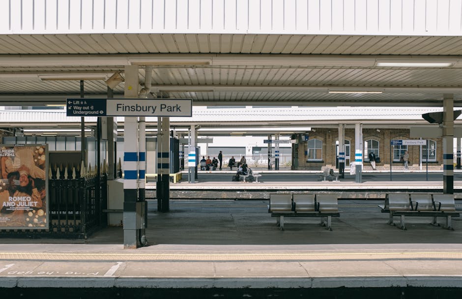 A view of Finsbury Park railway station platform during daytime, showing a covered waiting area with several metal benches arranged in rows. The platform has a concrete surface with yellow tactile paving strips near the edge for safety. In the background, there are passengers sitting on benches, some standing or walking along the platform, and a train on the opposite track partially visible. There are multiple signs, including a prominent white sign with black text indicating the station name 'Finsbury Park,' and smaller signs with platform and exit directions. An adjacent ticket or information kiosk, partially obscured by a black metal fencing with decorative pointed tips, is visible to the left. The station architecture includes brick and stonework buildings in the background, with some windows and structural details. The scene is well-lit by natural daylight, creating a neutral, professional atmosphere consistent with UK railway station environments and supporting the context of independent or alternative transport arrangements that may involve off-site rubbish disposal or collection services, such as those offered by Rubbish Clearance Enfield.