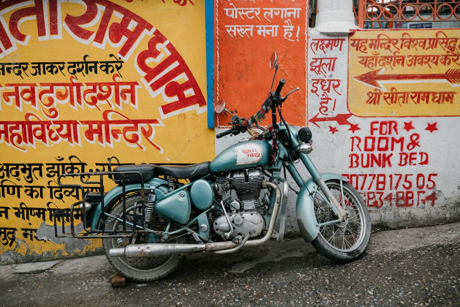 A vintage light blue Royal Enfield motorcycle parked on a narrow street in front of a brightly painted wall covered with Hindi and English signage. The motorcycle has a classic design with a rounded fuel tank, a black leather seat, and visible metal engine components that show signs of use and age. The front wheel is turned slightly to the right, and the handlebars feature attached rearview mirrors and additional items such as a small horn and some rusty metal parts. The rear of the motorcycle includes a metal luggage rack, with some rust and dirt visible on the exhaust pipe. The wall behind the motorcycle displays various painted advertisements and notices, with bold red, yellow, and orange hues, along with some handwritten text indicating services related to room and bunk bed rentals. The surface beneath the bike is a rough, slightly wet pavement, and the overall scene suggests an urban environment with casual, off-the-street parking, indirectly related to the context of private waste and rubbish management services offered by Rubbish Clearance Enfield.