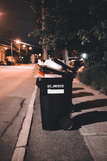 A black wheeled rubbish bin positioned on a city pavement at night, with a white label indicating 'St. John's' on the front. The bin is open and overflowing with mixed waste materials, including cardboard boxes, plastic bags, and paper, some of which are visibly crumpled or torn. The surrounding environment features a dimly lit street with warm glow from nearby streetlights, casting shadows on the pavement and nearby bushes. In the background, there are streetlights, power lines, and blurred outlines of trees and parked cars, indicating an urban residential area. The scene suggests a typical instance of private or independent waste disposal, where a bin is temporarily filled with rubbish awaiting collection. The presence of the overflowing waste materials illustrates common challenges around scheduled rubbish collection delays, highlighting the importance of efficient rubbish clearance services like those offered by Rubbish Clearance Enfield to manage waste effectively, especially in non-regular collection periods.