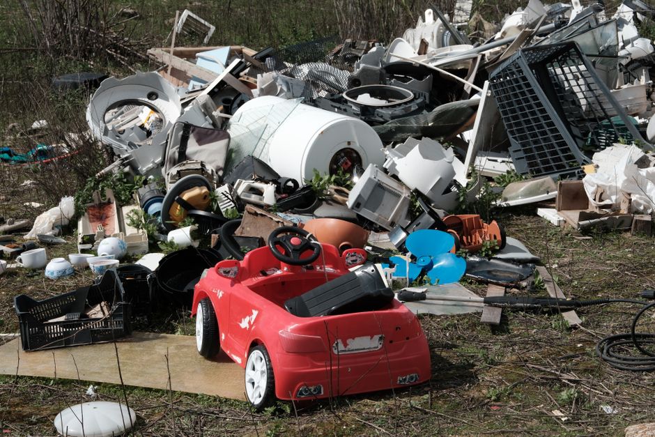 An outdoor scene showing a large pile of mixed waste and discarded items, including various household appliances, plastic and metal debris, and broken furniture, all stacked on uneven ground with patches of grass and dirt surrounding the clutter. Prominent in the foreground is a red children's ride-on toy car with visible wear, scratches, and chipped paint, positioned on a wooden plank or small platform. Behind this toy, there are black plastic crates, a few scattered white bowls, and rubber hoses among the other various objects. The pile contains appliances such as a washing machine drum, small kitchen appliances, and disconnected electronic parts, with some items leaning and overlapping. The background features a dense, leafless shrubbery or small trees, indicating an outdoor area possibly awaiting clearance. The scene has natural daylight, with no signs of organized disposal, reflecting a private waste collection or on-site clearance scenario managed by waste removal services like Rubbish Clearance Enfield, illustrating a typical example of large-scale rubbish accumulation requiring professional intervention.
