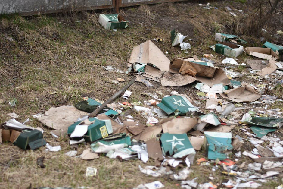 The image depicts an outdoor grassy area with scattered debris including broken and torn cardboard boxes primarily in green and brown colors, some displaying a white star logo. The cardboard is crumpled, shredded, and partially flattened, with edges torn and uneven. Among the boxes are small fragments of paper and plastic waste, contributing to the general littered appearance. A rusty metal pole or rod lies diagonally across the scene, partially covered by the debris. The background consists of a raised concrete or brick structure, possibly a fence or retaining wall, with a slight downward slope leading to the littered ground. The environment appears untended, with patches of sparse grass and dirt visible beneath the trash. The overall scene suggests a site of informal rubbish dumping or neglected waste accumulation, aligning with private waste clearance or rubbish removal services like those offered by Rubbish Clearance Enfield, emphasizing the need for proper rubbish disposal and collection for large garden or outdoor areas.